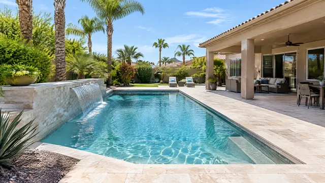 Upscale backyard pool at a 1990s-style stucco home in the Kierland area of northeast Phoenix: rectangular pool with a sheer descent water feature, light quartz finish, travertine decking, mature palms and a covered patio